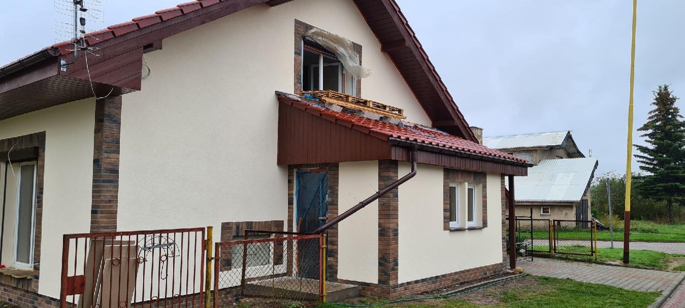 Dom jednorodzinny w trakcie remontu elewacji, z widocznymi rusztowaniami i zabezpieczeniami okien folią. Częściowo otynkowana ściana z dekoracyjnym wykończeniem.