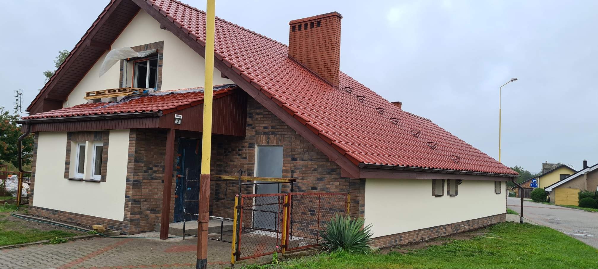 Dom jednorodzinny z czerwoną dachówką i elewacją w trakcie prac wykończeniowych. Widoczne rusztowania i zabezpieczone okno. Część elewacji pomalowana na kremowo.