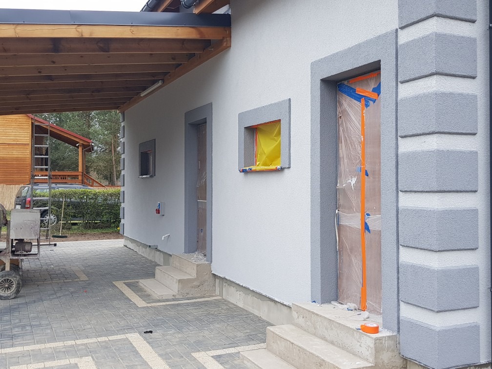 Elewacja domu w trakcie malowania: okna i drzwi zabezpieczone folią, świeżo pomalowane szare obramowania, widoczna faktura tynku i kostka brukowa na podjeździe.
