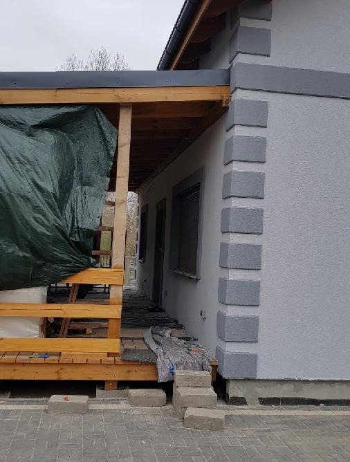 Szary dom z drewnianą konstrukcją zadaszenia, częściowo zakryty zieloną plandeką. Widoczne bloczki betonowe i fragment kostki brukowej.