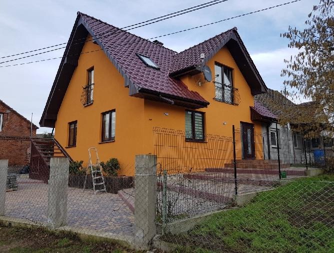 Dwupiętrowy dom z pomarańczową elewacją i ciemnoczerwonym dachem, widoczny zza ogrodzenia. Na ścianie drabina, a na dachu okno. Widoczne elementy wykończenia.