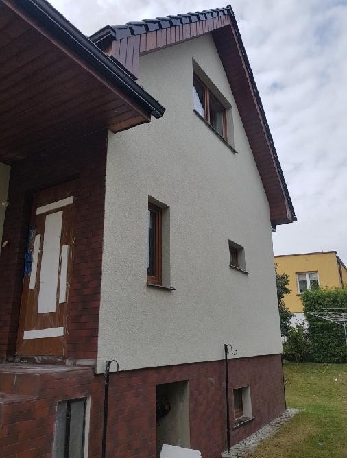Elewacja domu z oknami w brązowych ramach, jasnoszara ściana z fakturą, cokół z cegły klinkierowej. Widoczne schody wejściowe i fragment dachu z brązową dachówką.