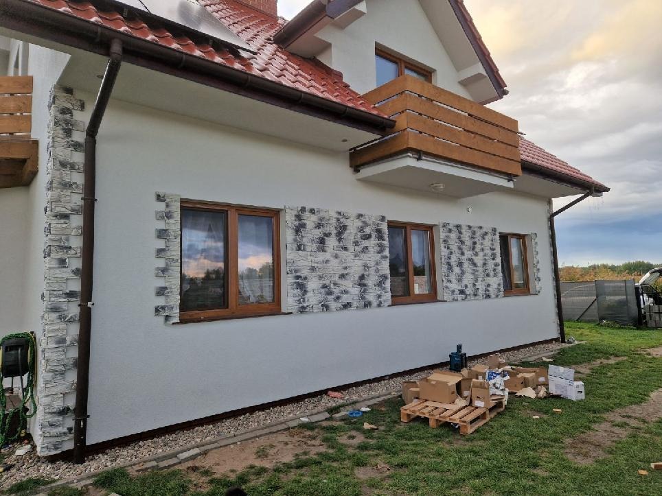 Elewacja domu z dekoracyjną okładziną kamienną wokół okien i na narożniku. Widoczne kartony i paleta na trawniku, sugerujące prace wykończeniowe.