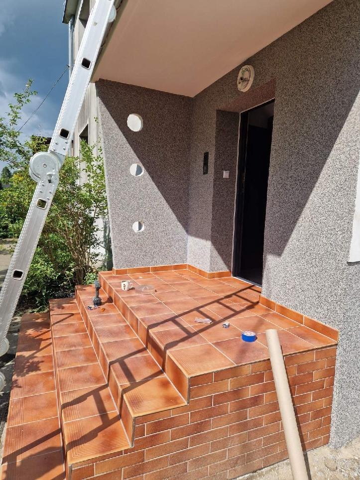 Wykończone płytkami schody wejściowe do domu z szarą elewacją. Widoczna drabina, narzędzia i materiały budowlane. Detal: okrągłe otwory w elewacji.