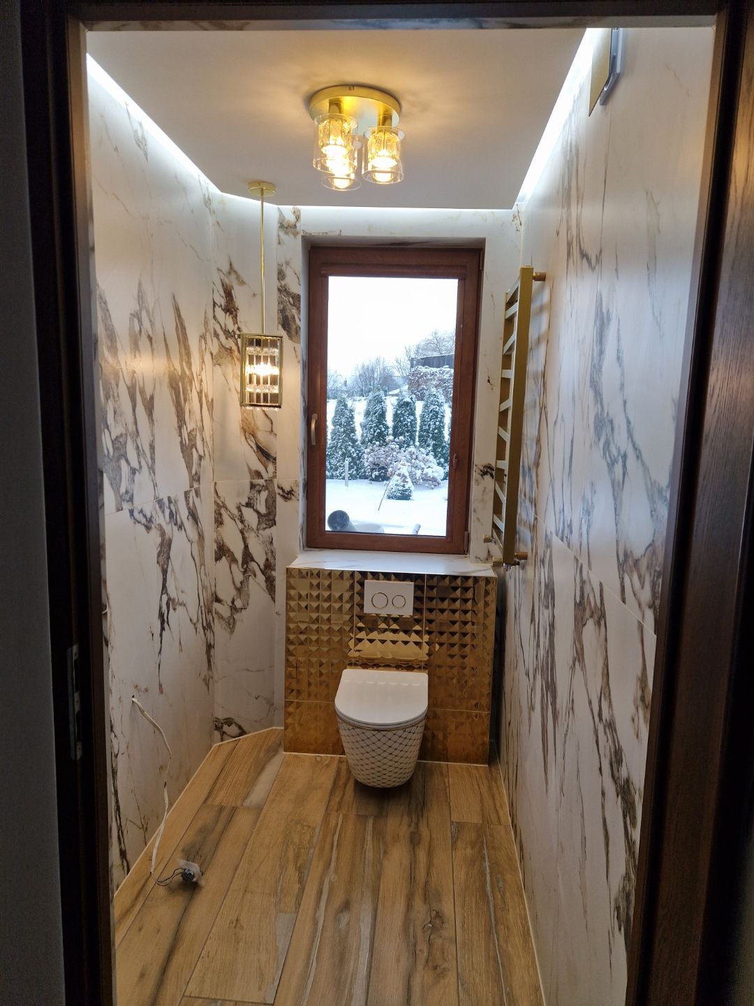 Elegancka toaleta z marmurowymi ścianami, drewnopodobną podłogą i złotymi akcentami. Okno z widokiem na zimowy krajobraz. Nowoczesne oświetlenie.