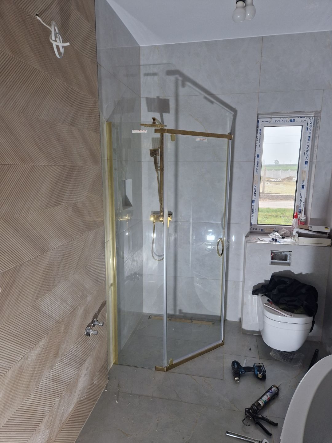 Nowoczesna kabina prysznicowa z złotymi detalami w łazience z szarą glazurą i narzędziami na podłodze. Widok na zewnątrz przez okno.