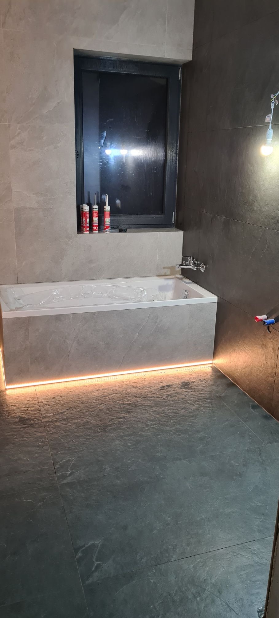 Nowoczesna łazienka z szarymi płytkami, podświetloną wanną i czarnym oknem. Widoczne silikony przy oknie. Minimalistyczny design, ciepłe oświetlenie LED.