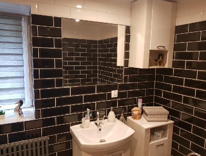 Nowoczesna łazienka z czarnymi, prostokątnymi płytkami i białymi fugami. Biała szafka z umywalką i lustro. Dekoracyjny akcent: figurka konika morskiego na parapecie.