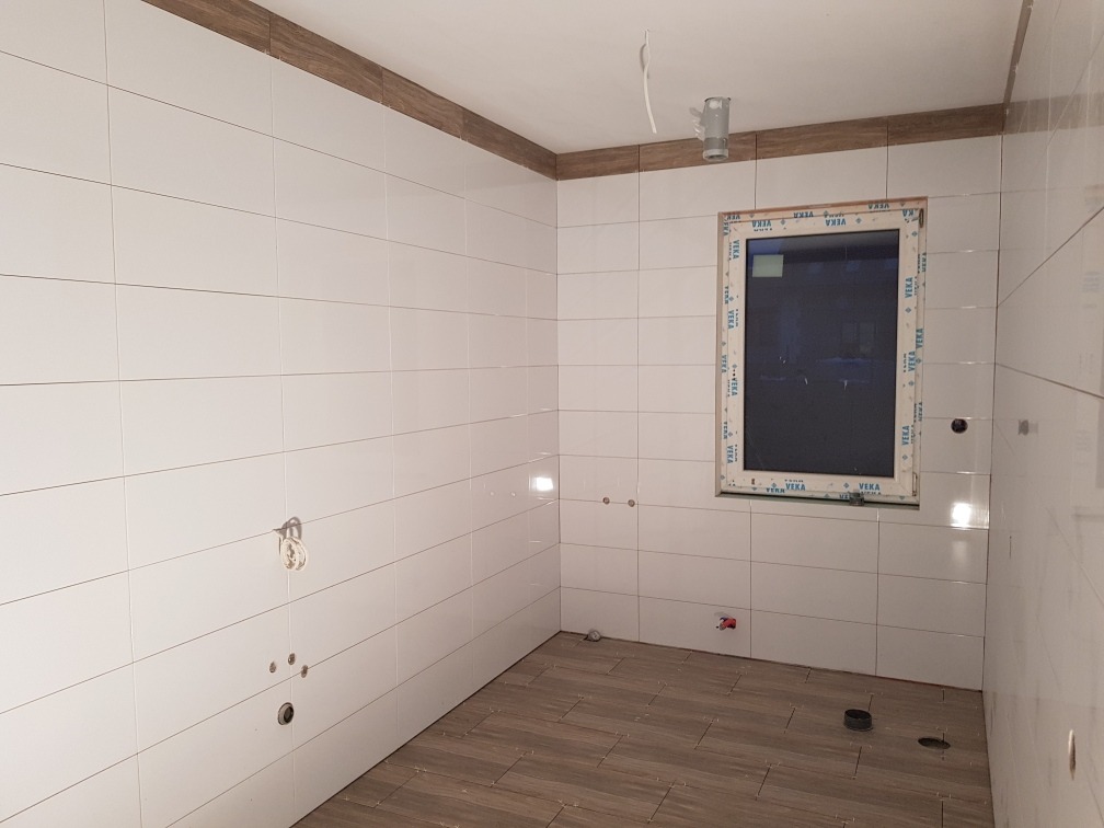 Wykończona kuchnia z białymi płytkami ściennymi i brązową listwą dekoracyjną. Na podłodze brązowe płytki imitujące drewno. Okno z plastikową ramą.