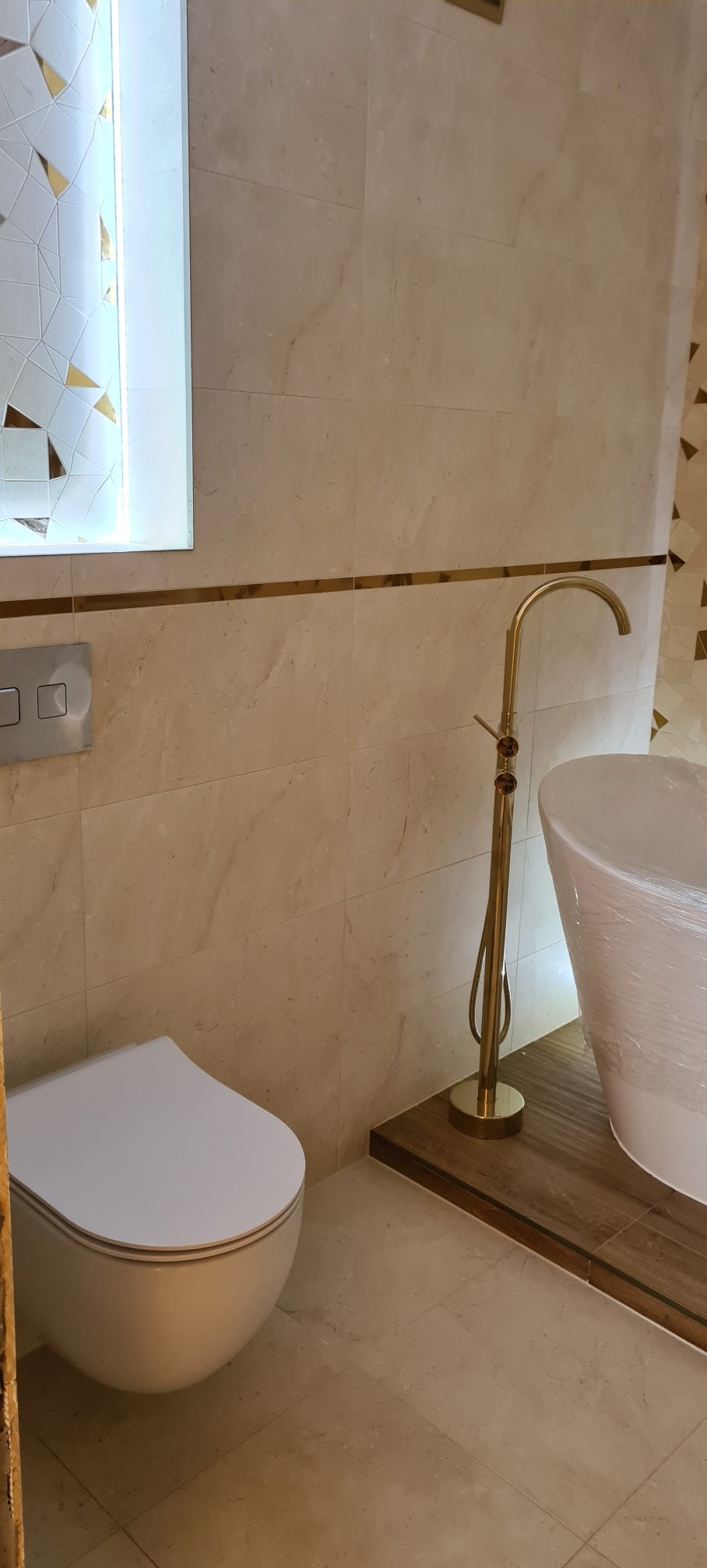 Elegancka łazienka w beżowych płytkach z złotymi akcentami: bateria wolnostojąca, podświetlane lustro z mozaiką i podwieszana toaleta.