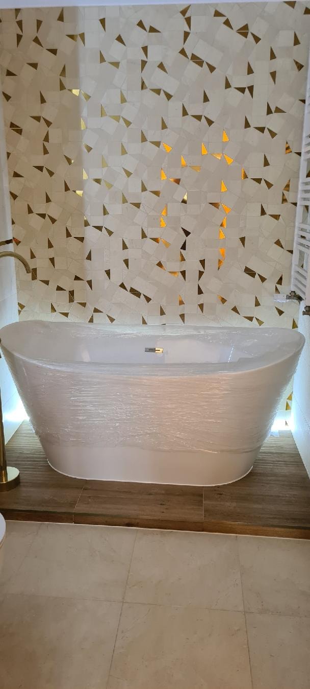 Nowoczesna łazienka z wolnostojącą wanną owiniętą folią ochronną, drewnopodobną podłogą i mozaiką ścienną z złotymi akcentami. Oświetlenie LED podkreśla detale.