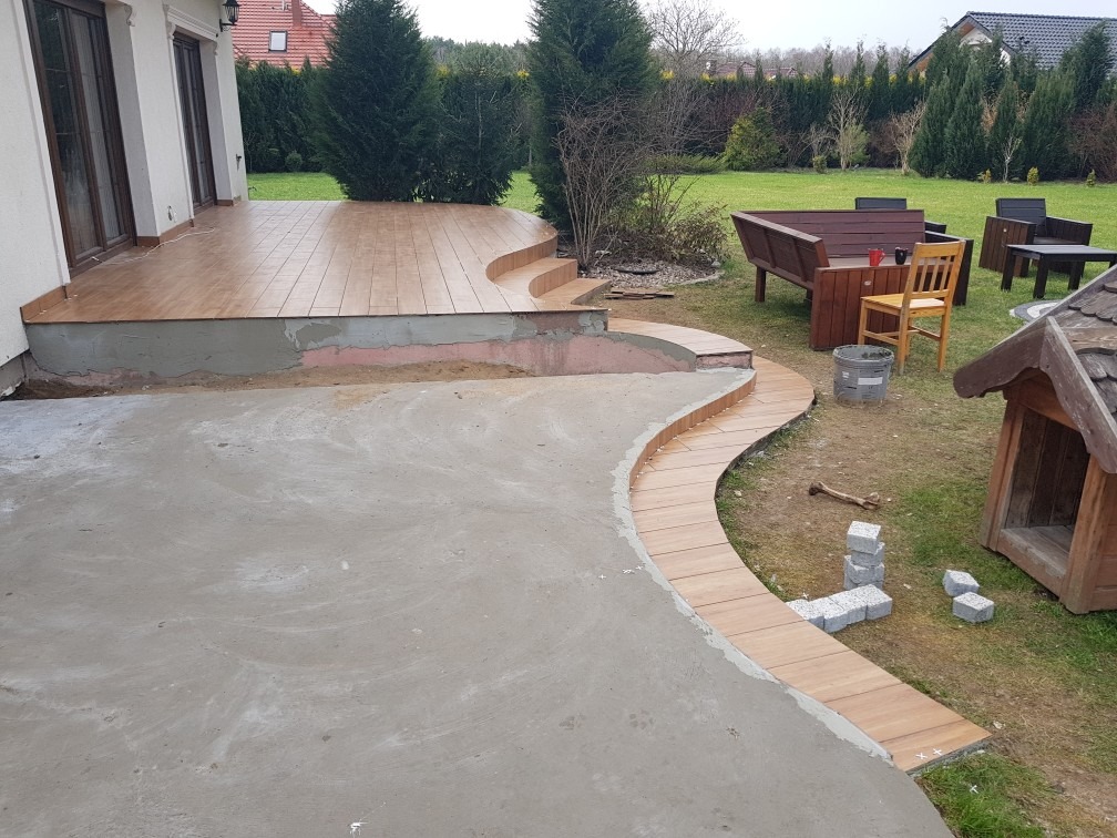 Układanie płytek imitujących drewno na tarasie i ścieżce w ogrodzie. Częściowo ukończone, widoczne materiały i narzędzia. Tło: dom jednorodzinny i zieleń.