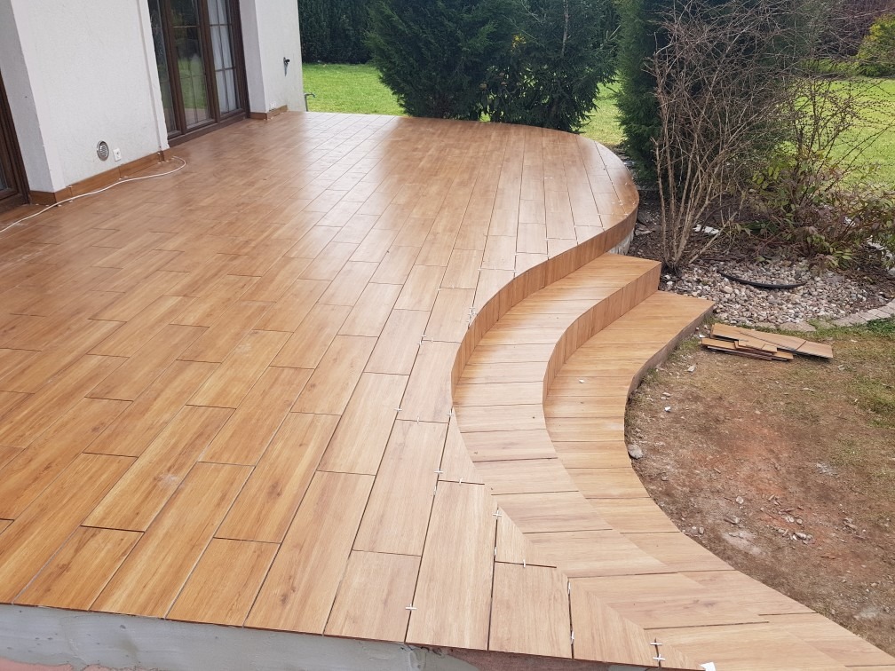 Balkon z drewnopodobną terakotą ułożoną na podłodze i schodach, widok z góry, jasne oświetlenie, tło z zielenią.