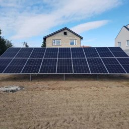 THERMOENERGY SPÓŁKA Z OGRANICZONĄ ODPOWIEDZIALNOŚCIĄ - Instalacja fotowoltaiczna na gruncie, panele w rzędach na metalowej konstrukcji, w tle domy jednorodzinne i błękitne niebo z chmurami.