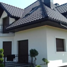 THERMOENERGY SPÓŁKA Z OGRANICZONĄ ODPOWIEDZIALNOŚCIĄ - Biały dom z ciemną dachówką i balkonami z kutą balustradą. Pracownik na drabinie przy wejściu. Widoczne okna z roletami zewnętrznymi i ozdobne krzewy.