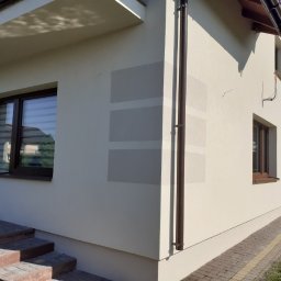 THERMOENERGY SPÓŁKA Z OGRANICZONĄ ODPOWIEDZIALNOŚCIĄ - Jasna elewacja domu jednorodzinnego z widocznymi fragmentami siatki podtynkowej, brązowe okna i rynna. Ujęcie z poziomu gruntu, widoczne schody wejściowe.