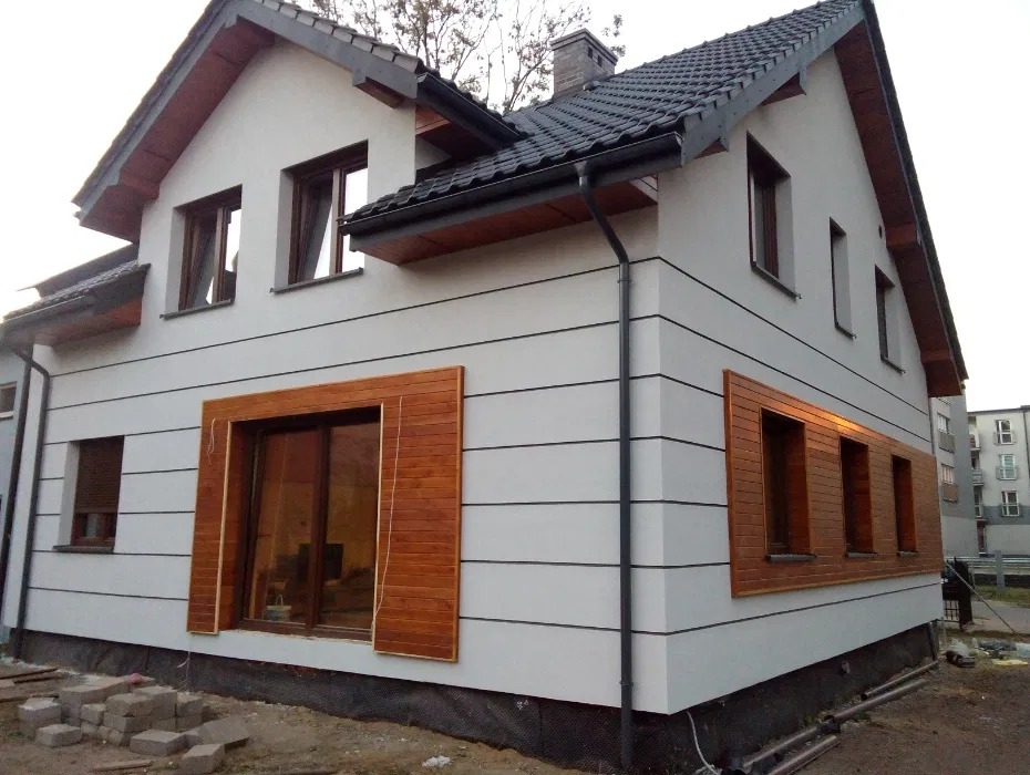 Biała elewacja domu z drewnianymi panelami okiennymi i ciemną dachówką. Widoczne detale wykończenia i elementy konstrukcyjne. Ujęcie z zewnątrz.