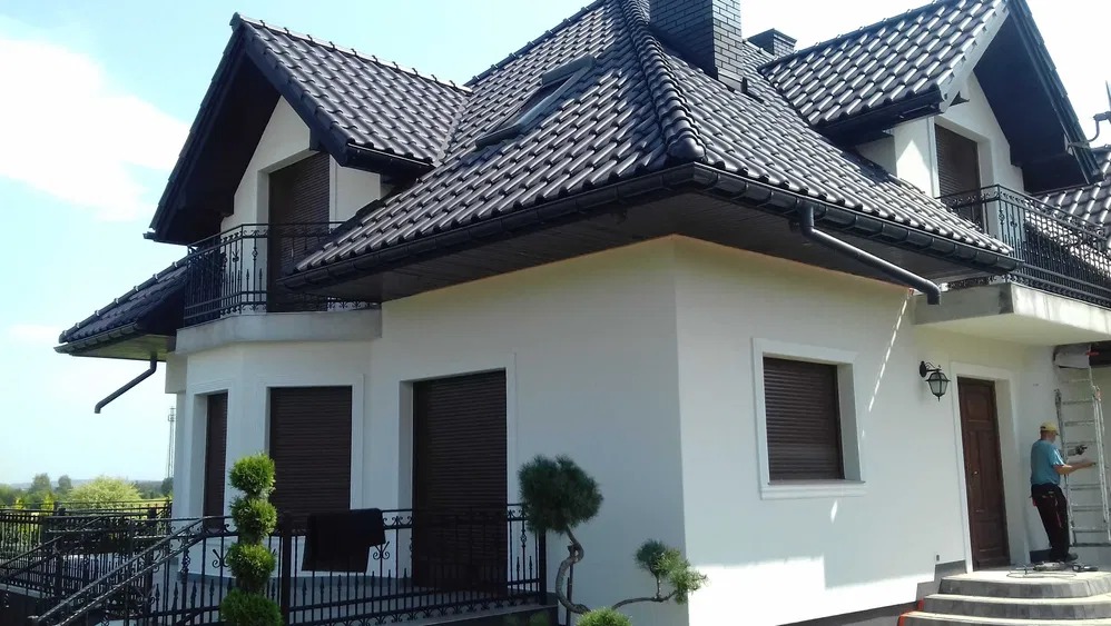 Biały dom z ciemną dachówką i balkonami z kutą balustradą. Pracownik na drabinie przy wejściu. Widoczne okna z roletami zewnętrznymi i ozdobne krzewy.
