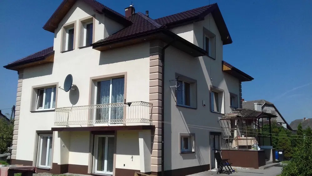 Dwupiętrowy dom z beżową elewacją, brązowym dachem i białymi oknami. Widoczny balkon z metalową balustradą i satelita na ścianie. Słoneczny dzień.