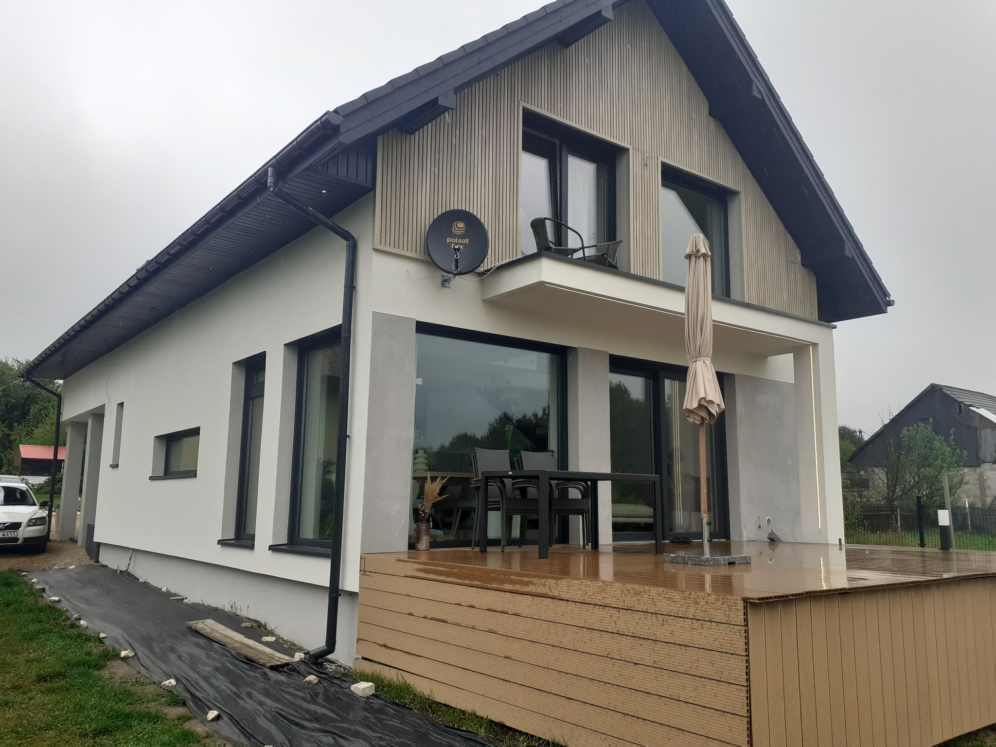 Nowoczesny dom z białą elewacją i drewnianą okładziną na poddaszu. Widoczny taras z meblami ogrodowymi i satelita na ścianie. Minimalistyczny design, pochmurna pogoda.