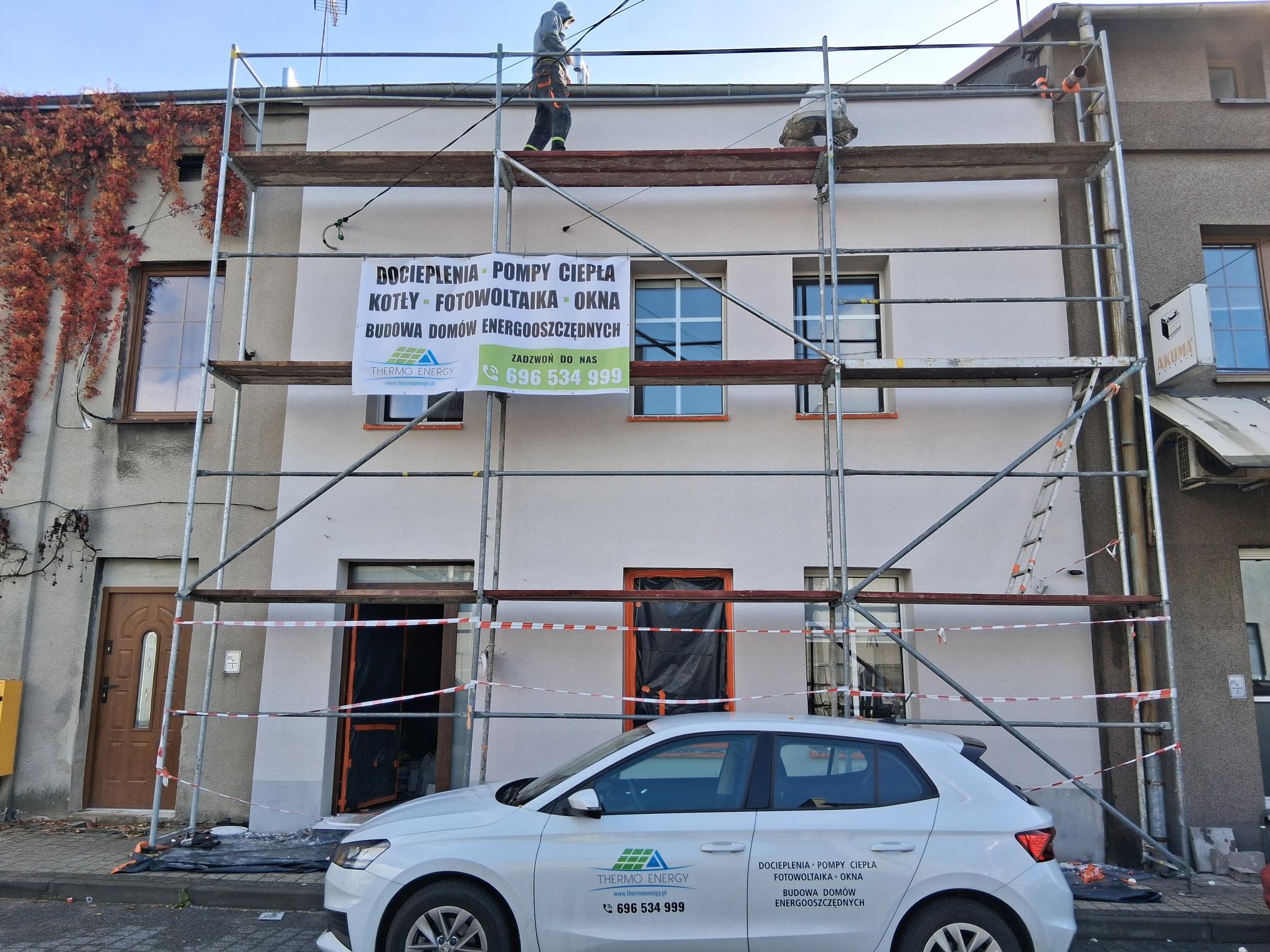 Elewacja budynku w trakcie prac: rusztowanie, pracownik na górze, baner reklamowy z ofertą dociepleń, pomp ciepła, fotowoltaiki i okien. Samochód firmowy z logo.