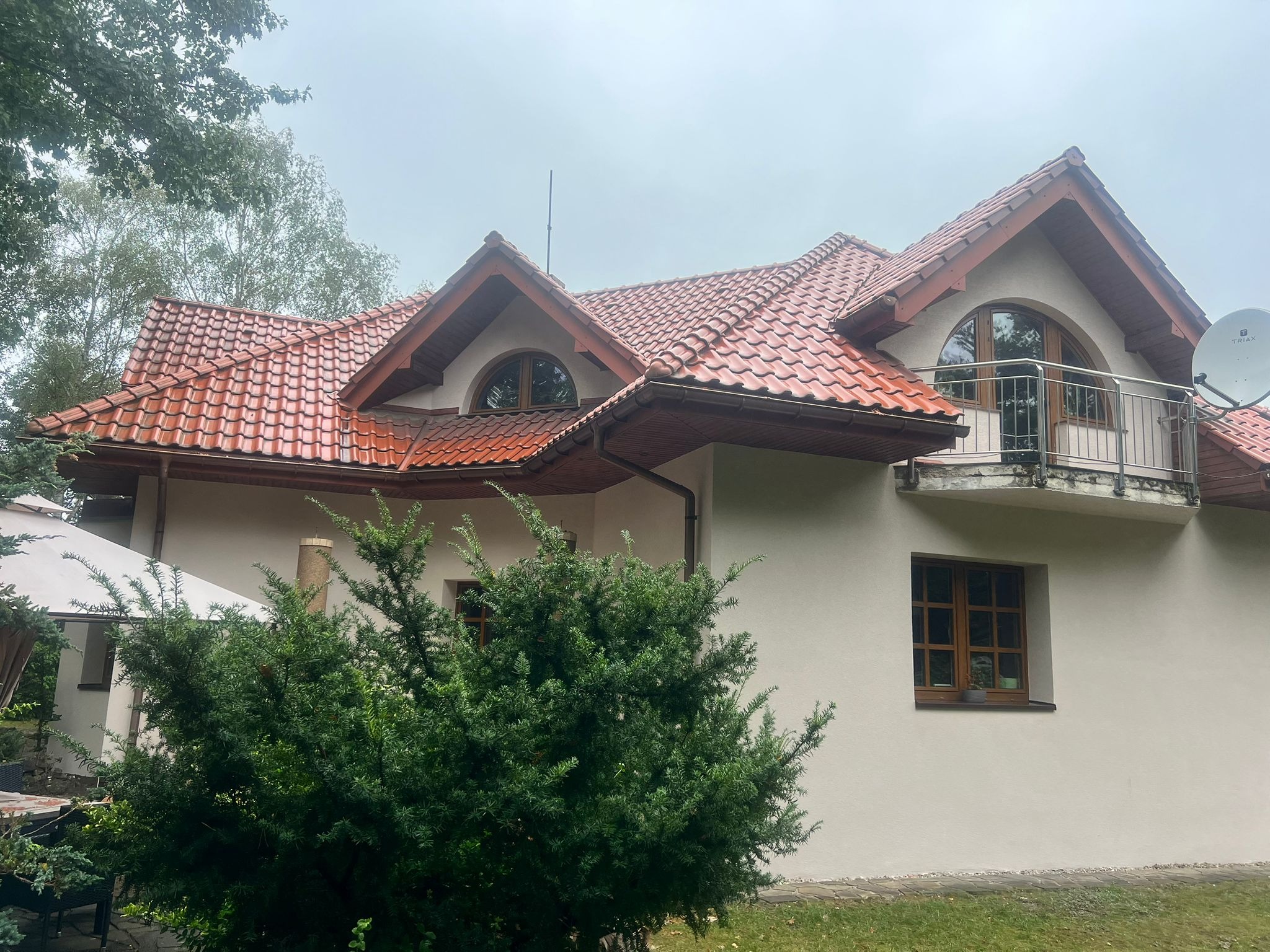 Jasny dom z czerwoną dachówką, balkonem i oknami z drewnianymi ramami. Na pierwszym planie zielony krzew. Widok na elewację i dach w pochmurny dzień.