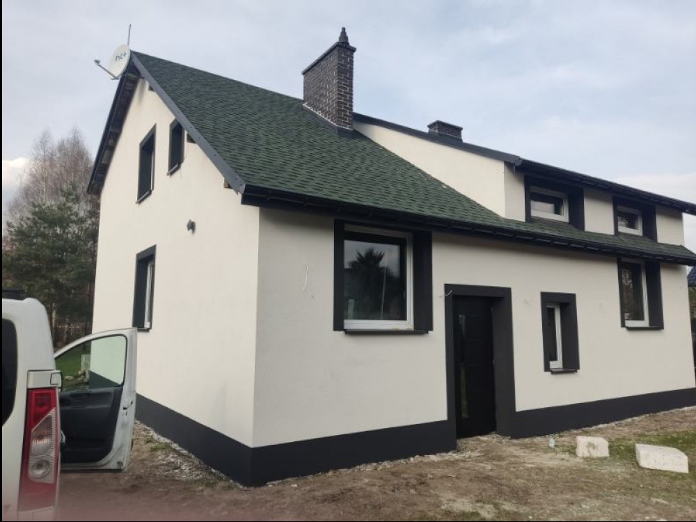 Otynkowany dom jednorodzinny z ciemną podmurówką i stolarką okienną. Zielony dach z kominem. Częściowo widoczny samochód dostawczy z otwartymi drzwiami.