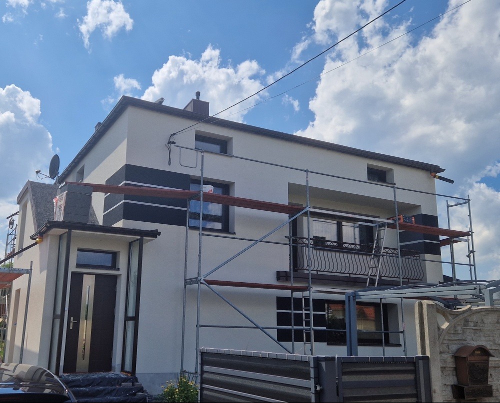 Dwukondygnacyjny dom z elewacją w odcieniach bieli i grafitu, częściowo osłonięty rusztowaniem. Widoczne okna, balkon z ozdobną balustradą i błękitne niebo w tle.