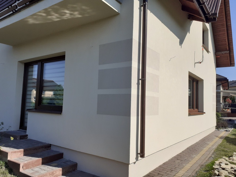 Jasna elewacja domu jednorodzinnego z widocznymi fragmentami siatki podtynkowej, brązowe okna i rynna. Ujęcie z poziomu gruntu, widoczne schody wejściowe.