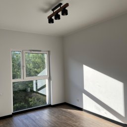 Robert Plewka RONERGY - Jasne wnętrze z drewnianą podłogą i oknem z widokiem na zieleń. Na suficie zamontowana lampa z trzema reflektorami. Promienie słoneczne oświetlają ścianę.