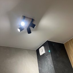 Robert Plewka RONERGY - Czarna, potrójna lampa sufitowa z drewnianą podstawą oświetla nowoczesną łazienkę z szarymi i czarnymi płytkami oraz białym włącznikiem wentylatora.