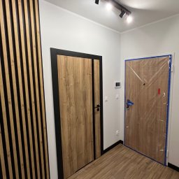 Robert Plewka RONERGY - Nowoczesne wnętrze: drewniane lamele na ścianie, drzwi z czarną ramą i oświetlenie szynowe z reflektorami. Jedne drzwi zabezpieczone folią malarską.