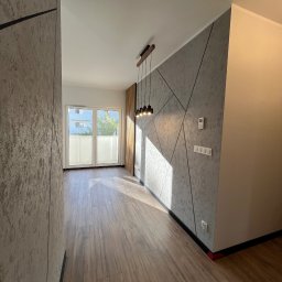 Robert Plewka RONERGY - Nowoczesne wnętrze z drewnianą podłogą, geometryczną ścianą i wiszącą lampą. Widoczne gniazdka elektryczne i termostat na ścianie.