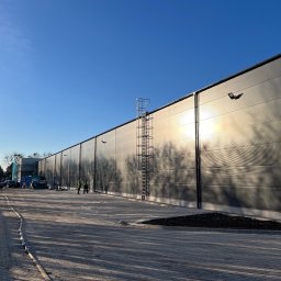 Robert Plewka RONERGY - Długa, szara ściana hali przemysłowej z zamontowanymi lampami i drabiną ewakuacyjną. W tle widoczni pracownicy w kamizelkach odblaskowych. Słoneczny dzień.