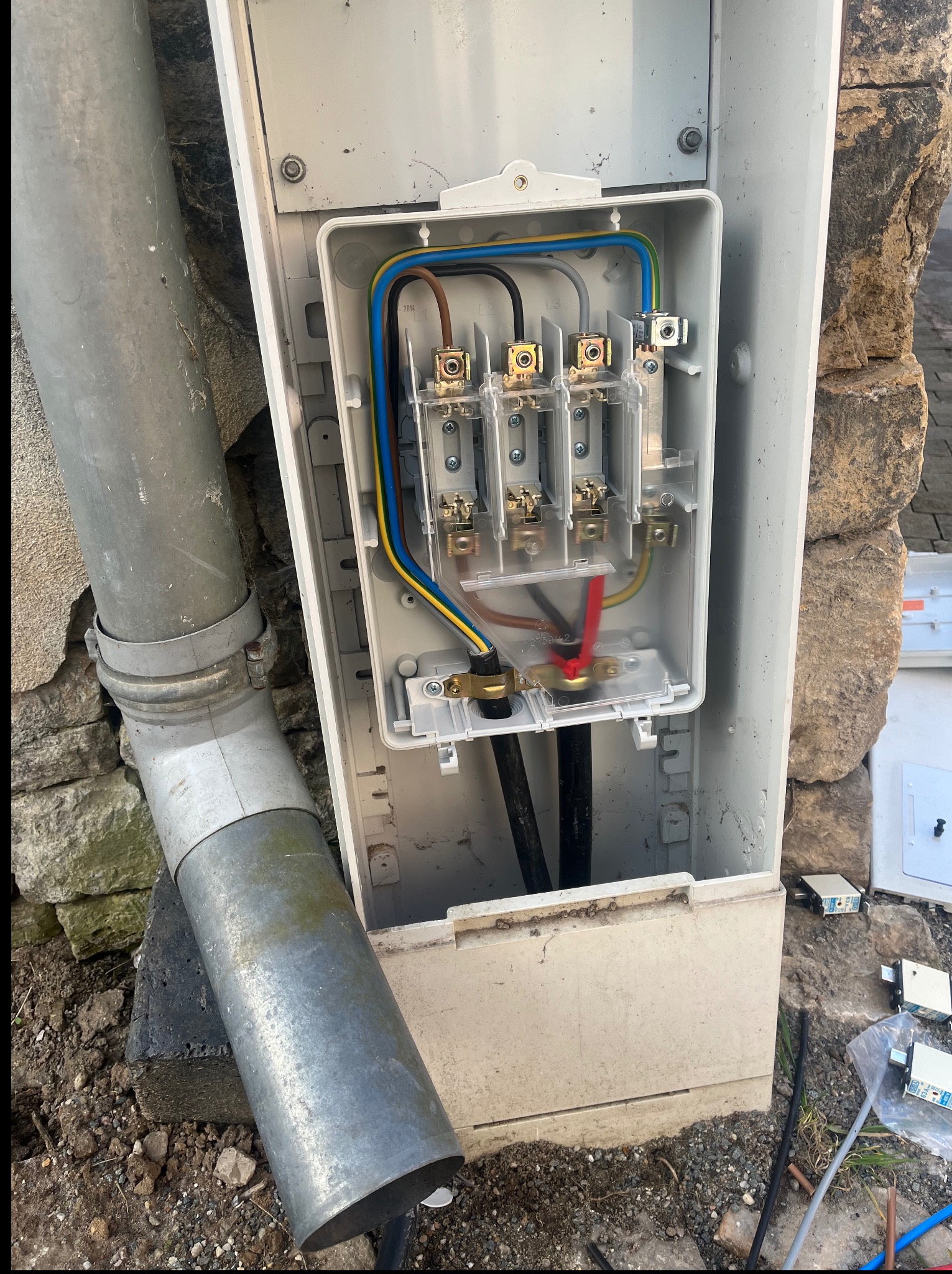 Otwarta skrzynka z bezpiecznikami i przewodami na tle kamiennej ściany. Widoczne elementy instalacji elektrycznej i rura osłonowa. Detal przyłącza.