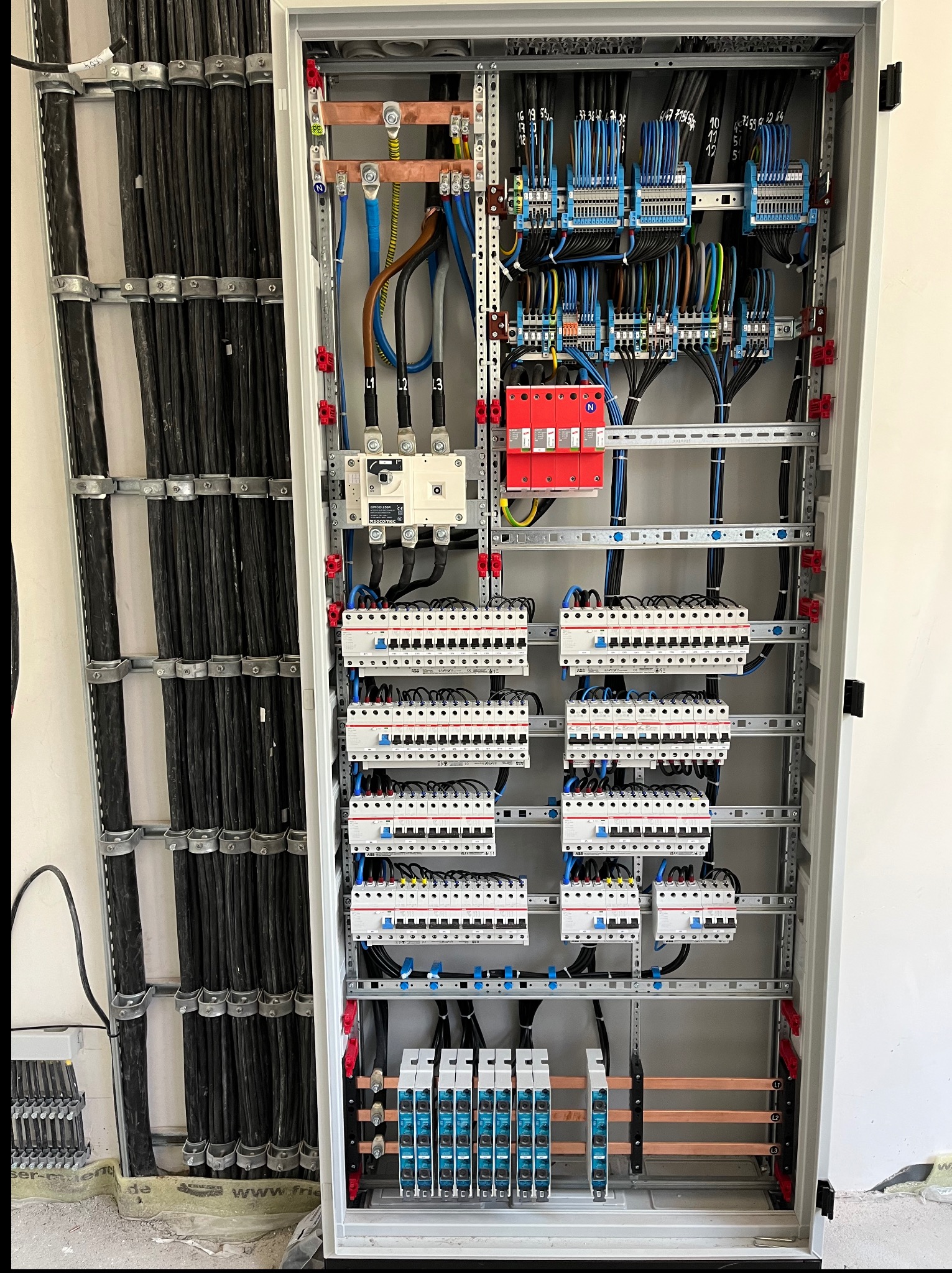 Rozdzielnica elektryczna z licznymi zabezpieczeniami, przewodami i szynami, zamontowana wewnątrz budynku. Widoczne kable prowadzone wzdłuż ściany.