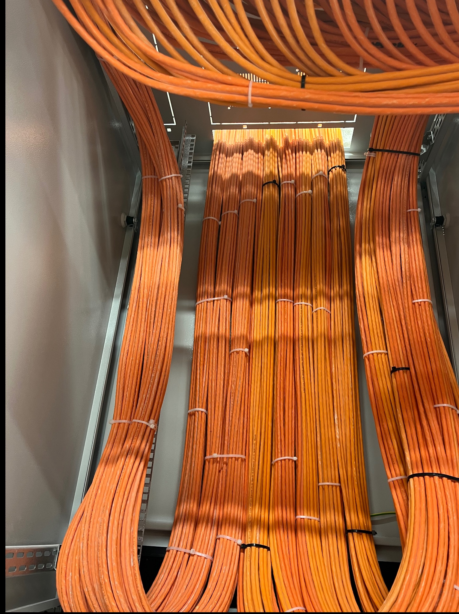 Pomarańczowe kable elektryczne ułożone równolegle w szafie sterowniczej, spięte opaskami. Widok z dołu, podkreślający porządek i profesjonalizm instalacji.