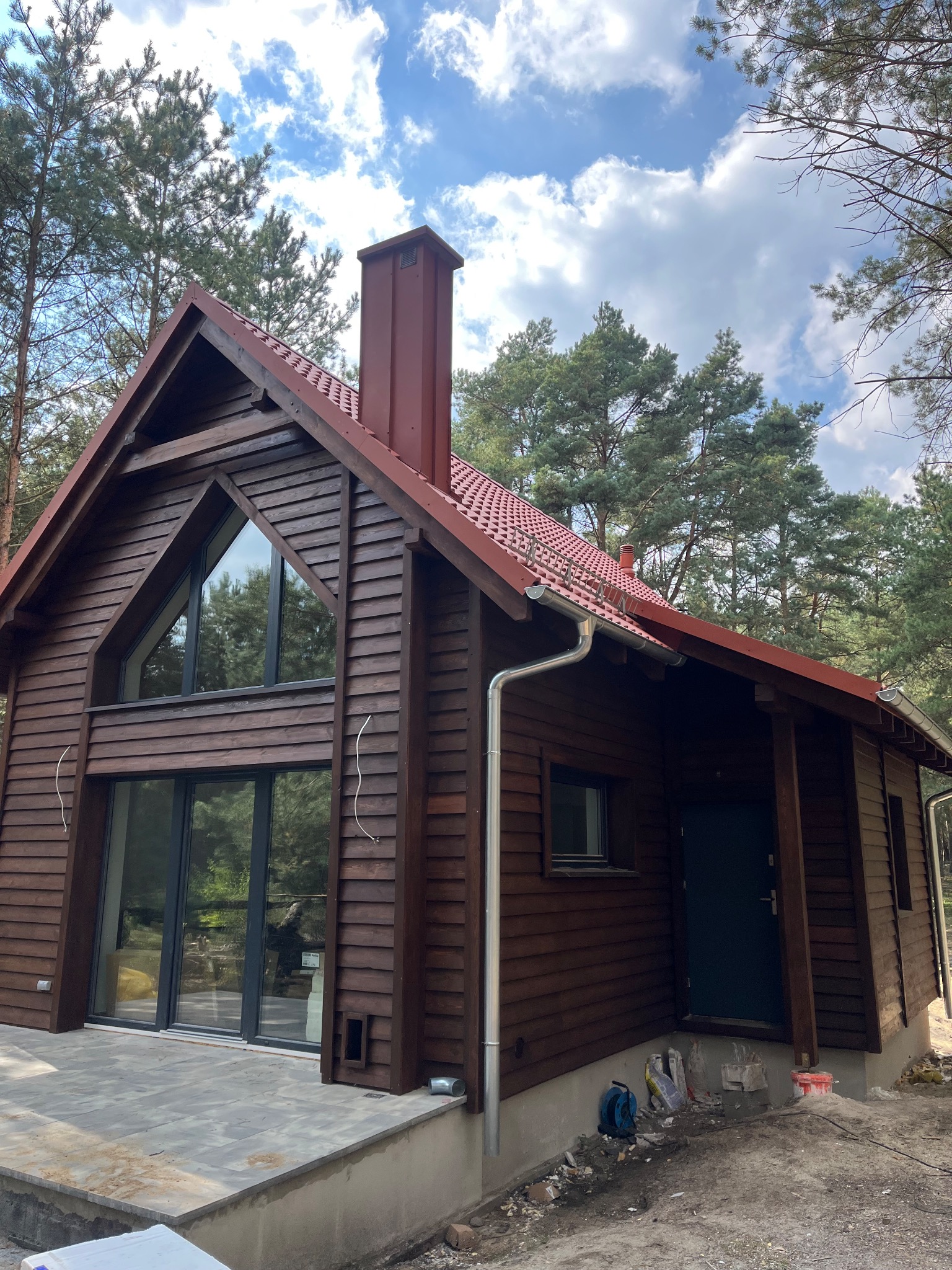 Drewniany dom z czerwoną dachówką i dużymi oknami, otoczony drzewami. Widoczne rynny i elementy instalacji na elewacji. Częściowo wykończony taras.