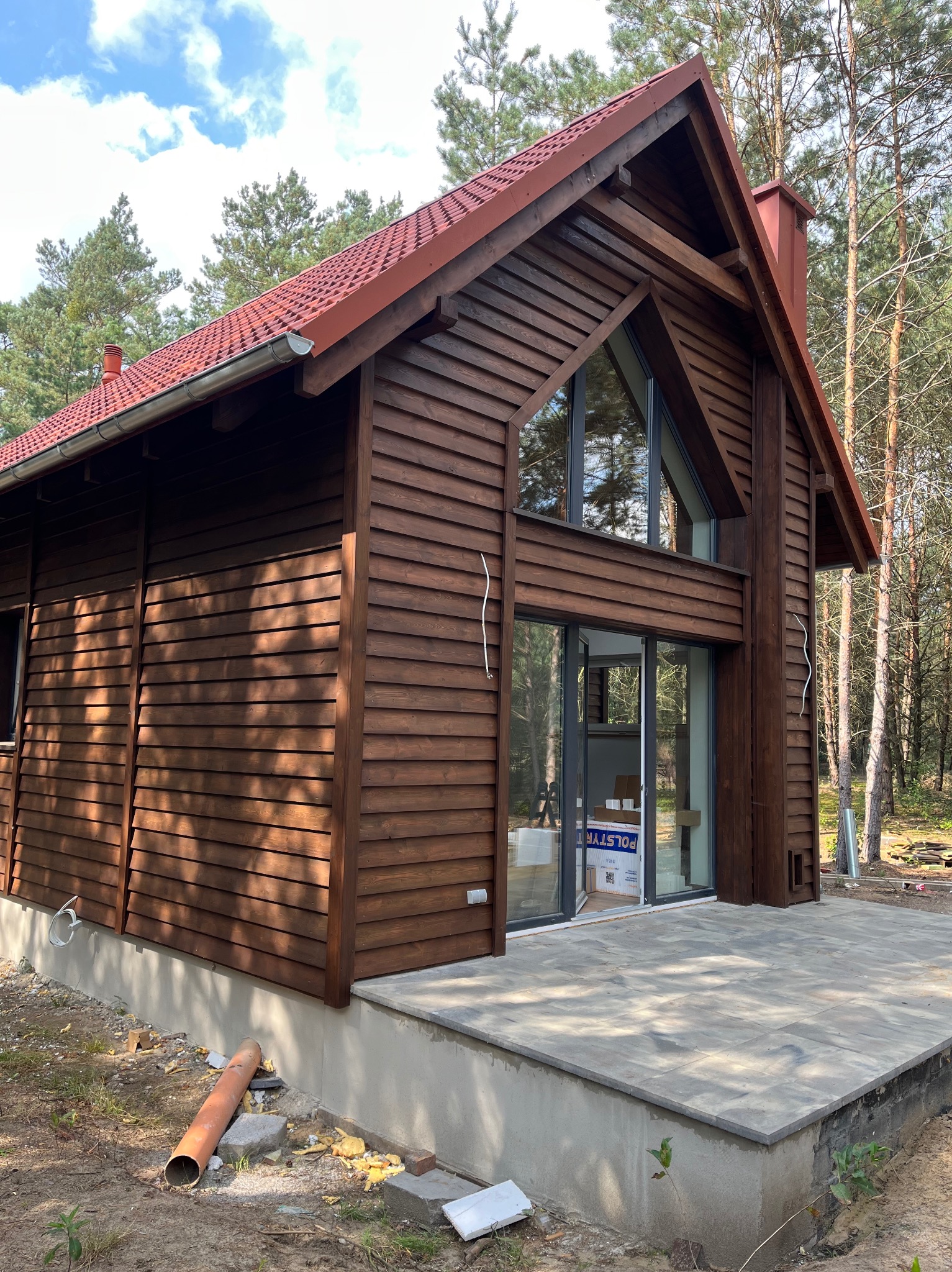 Nowoczesny, drewniany dom z czerwoną dachówką i dużymi oknami, otoczony lasem. Widoczny taras z szarych płyt i fragment instalacji na zewnątrz.