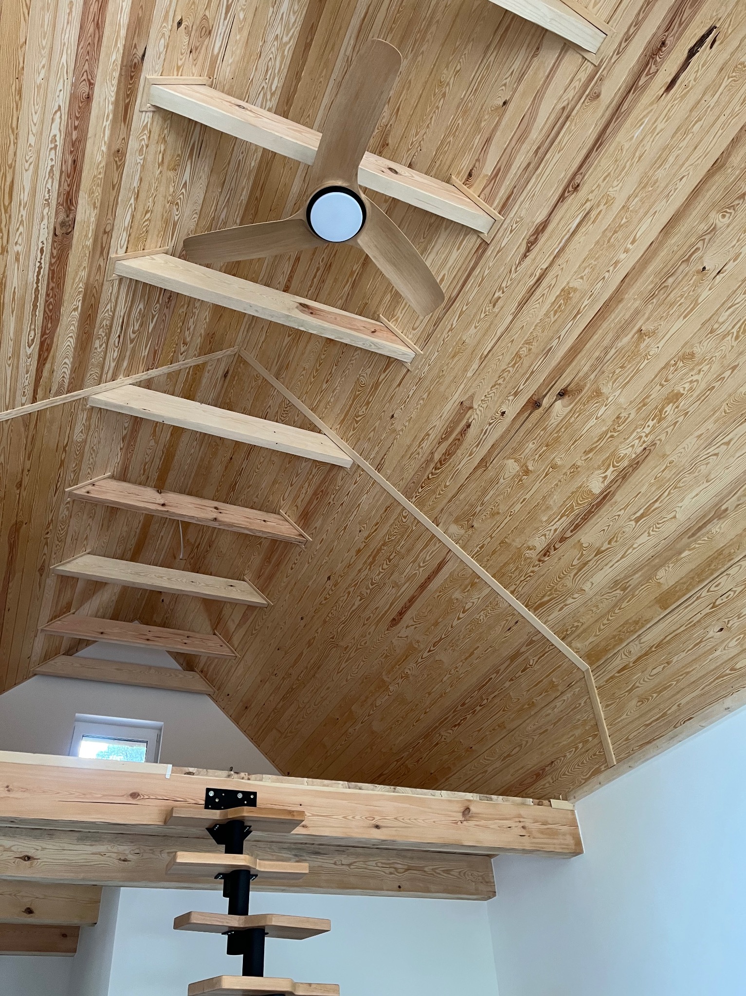 Drewniany sufit z widoczną konstrukcją i zamontowanym wentylatorem sufitowym z oświetleniem, widziany pod kątem. Naturalne wykończenie drewna.