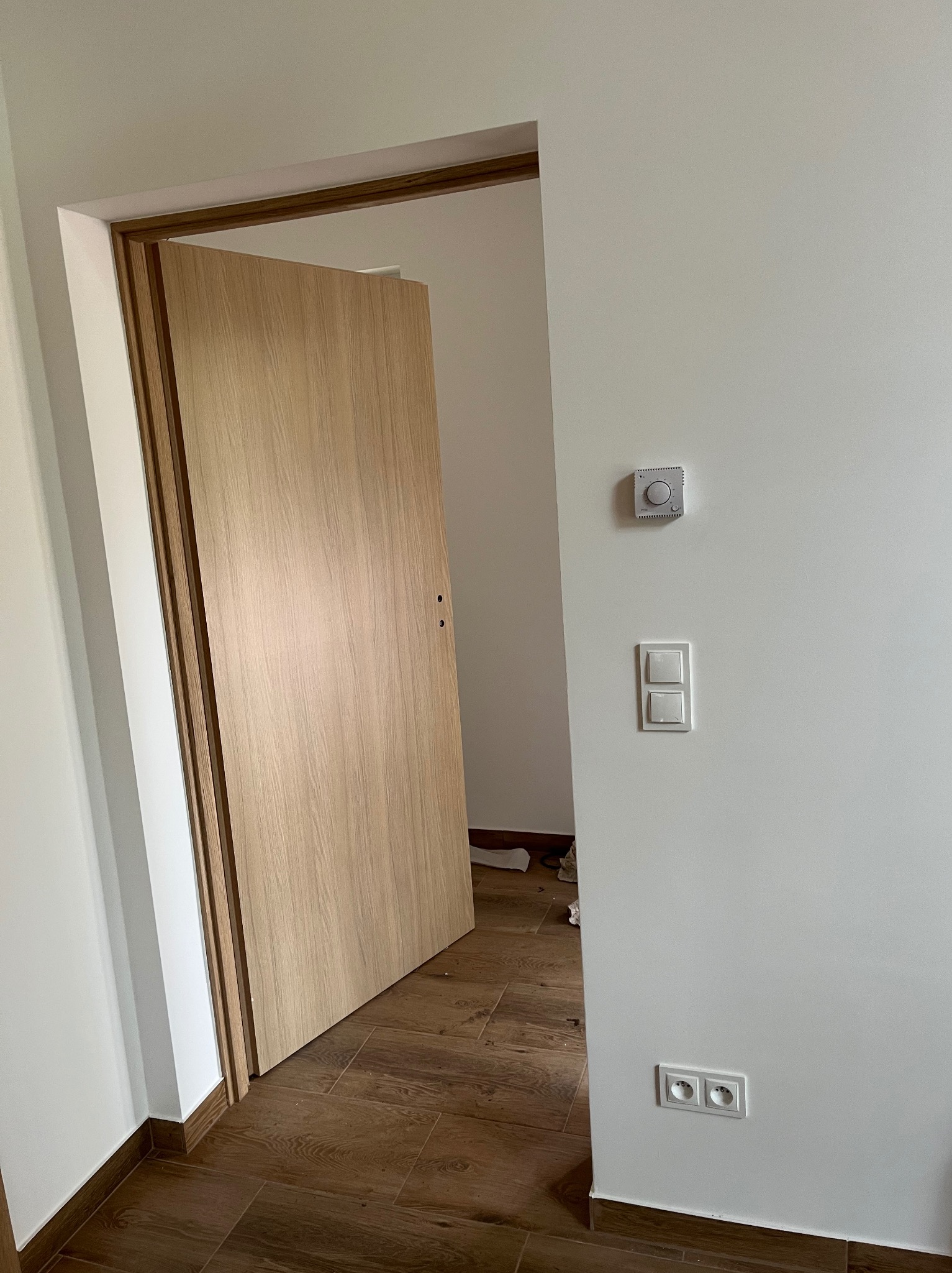 Jasne, drewniane drzwi wewnętrzne z ościeżnicą, widoczne gniazdka elektryczne i regulator temperatury na białej ścianie. Podłoga wyłożona brązowymi płytkami.