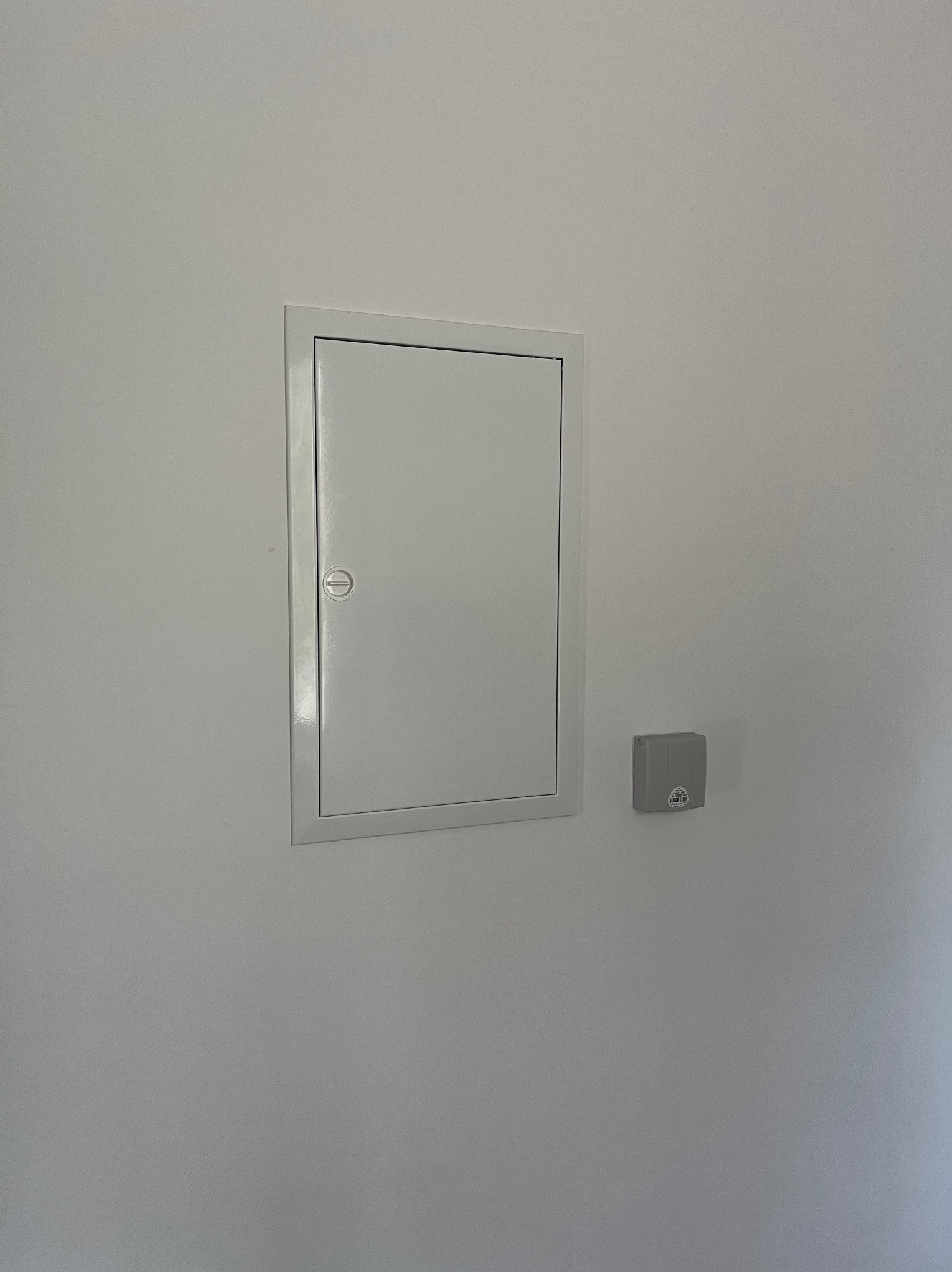 Szara, metalowa skrzynka elektryczna z zamkiem, wbudowana w ścianę, obok szara puszka instalacyjna. Minimalistyczny design, neutralne tło.