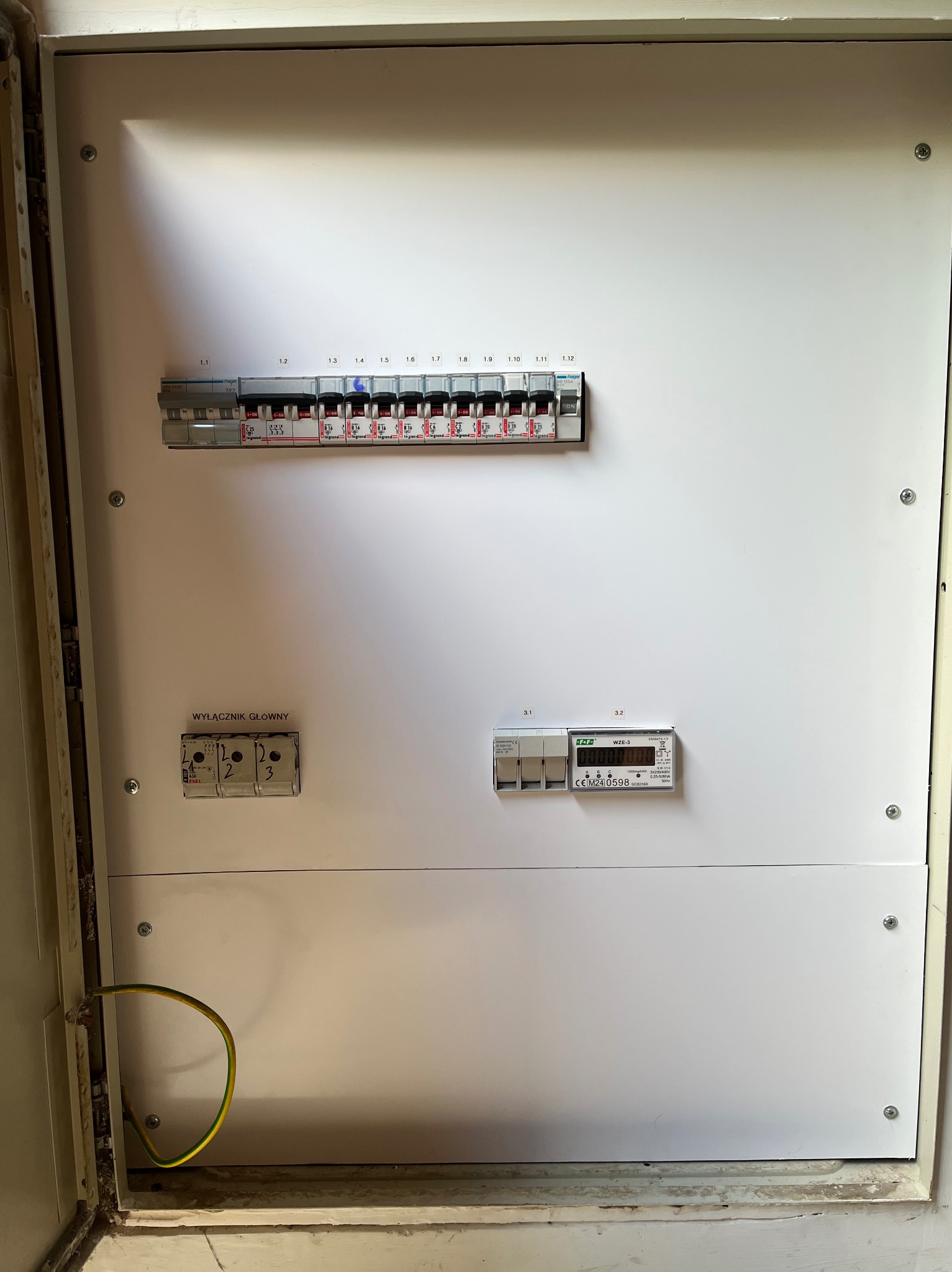 Biała skrzynka elektryczna z bezpiecznikami, wyłącznikiem głównym i licznikiem energii. Widoczny przewód uziemiający. Element instalacji elektrycznej.