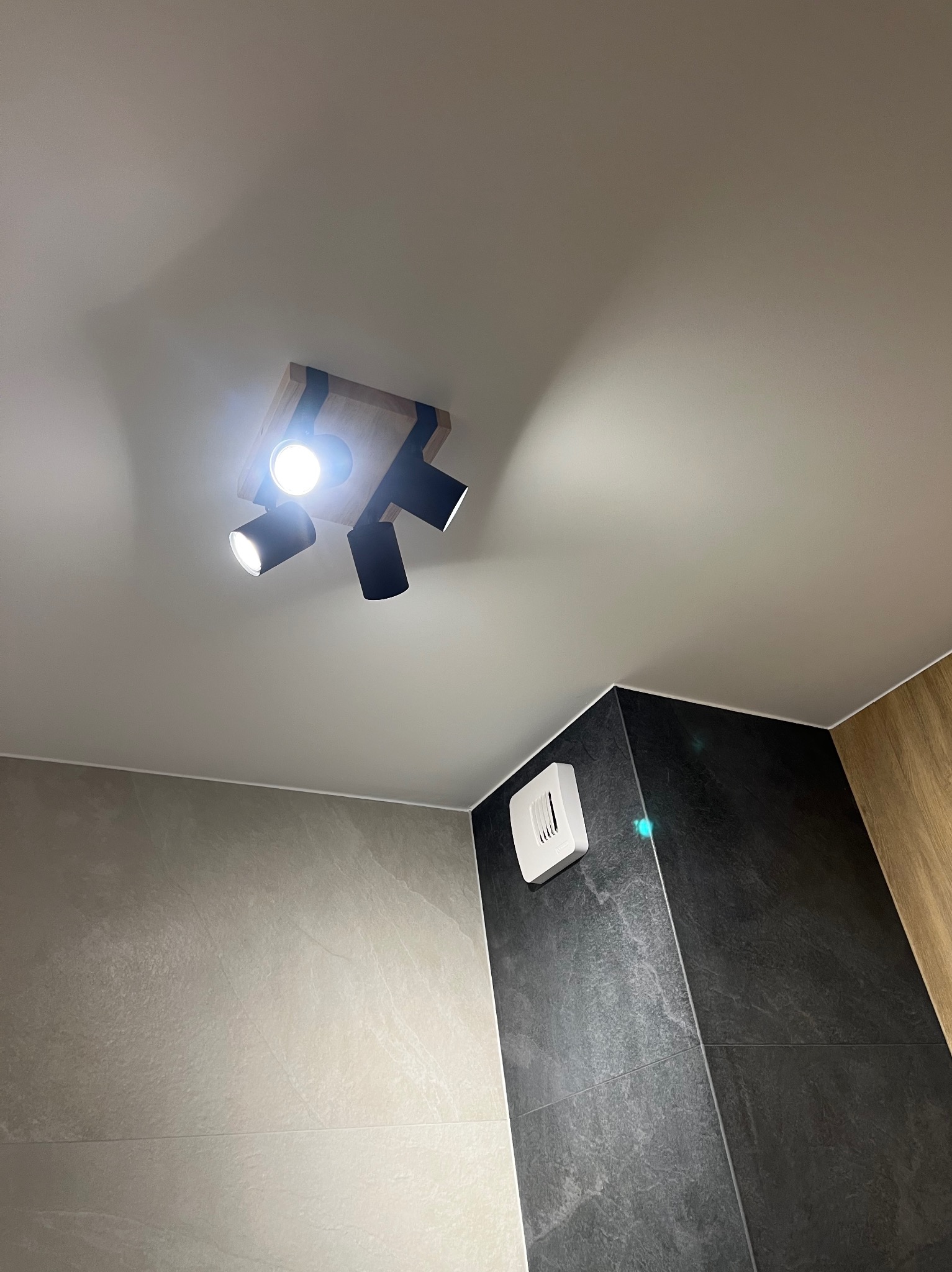 Czarna, potrójna lampa sufitowa z drewnianą podstawą oświetla nowoczesną łazienkę z szarymi i czarnymi płytkami oraz białym włącznikiem wentylatora.