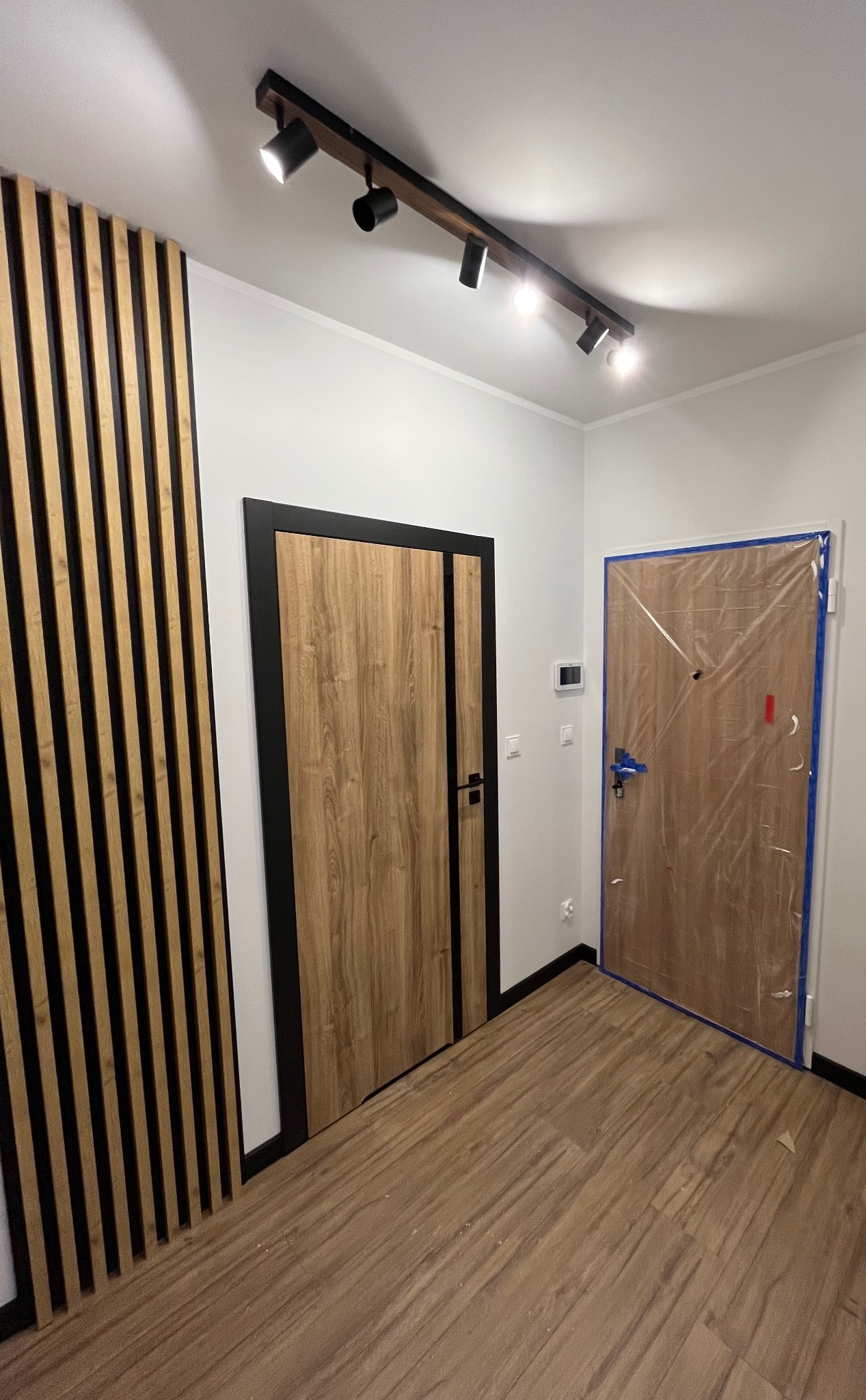 Nowoczesne wnętrze: drewniane lamele na ścianie, drzwi z czarną ramą i oświetlenie szynowe z reflektorami. Jedne drzwi zabezpieczone folią malarską.