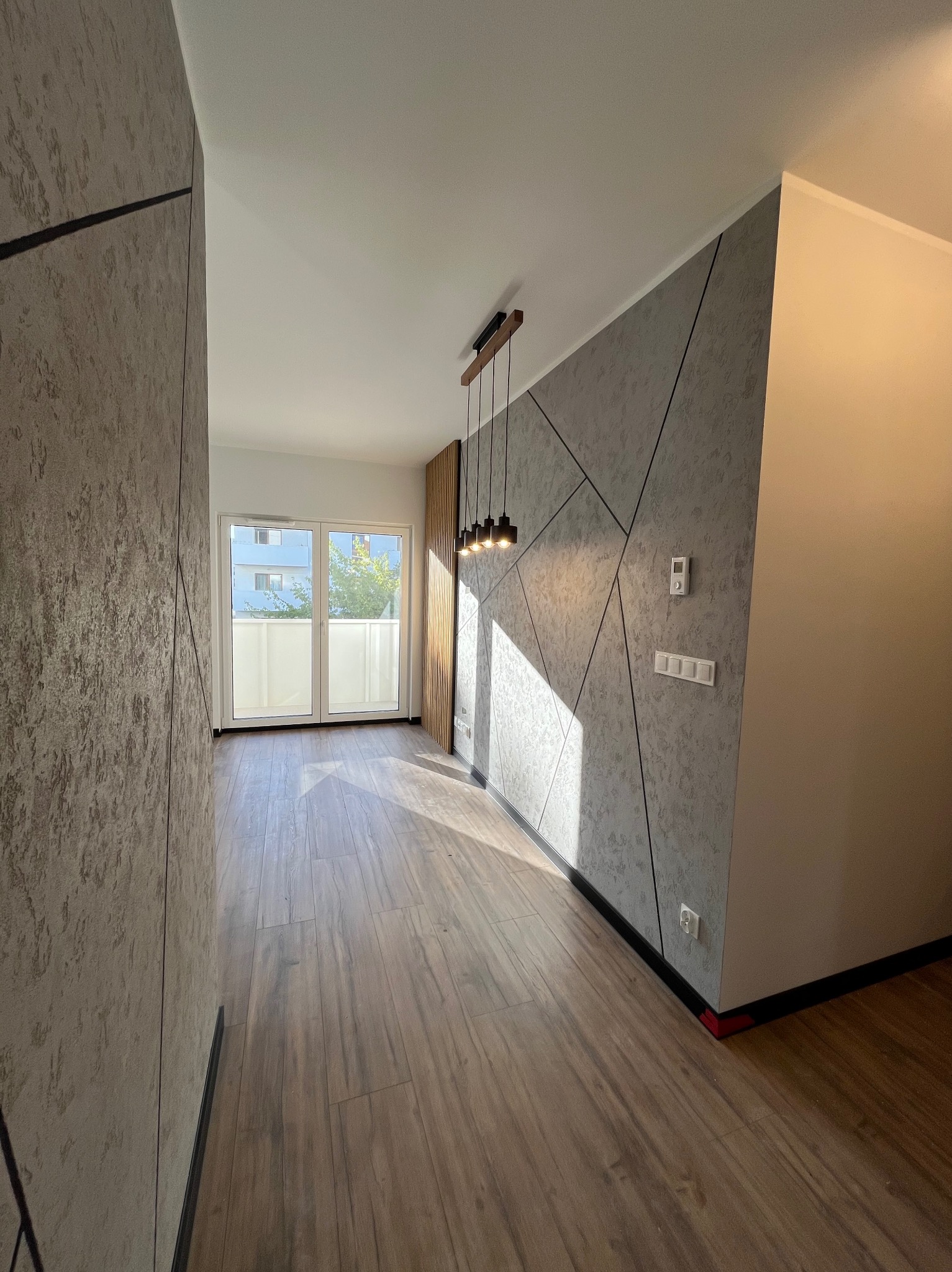 Nowoczesne wnętrze z drewnianą podłogą, geometryczną ścianą i wiszącą lampą. Widoczne gniazdka elektryczne i termostat na ścianie.