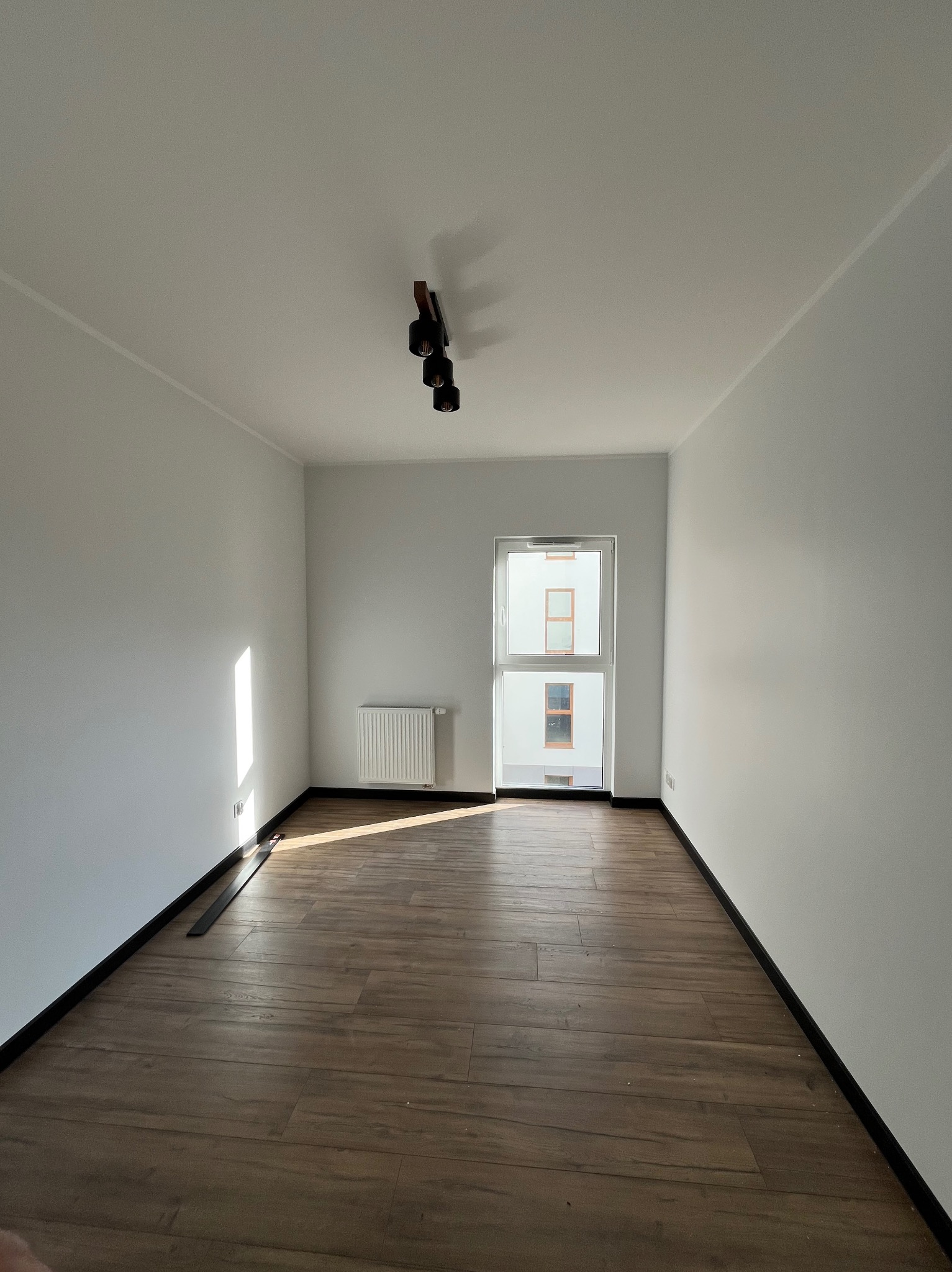 Pusty pokój z drewnianą podłogą, białymi ścianami i nowoczesną lampą sufitową. Widoczne okno z widokiem na budynek. Minimalistyczne wykończenie wnętrza.