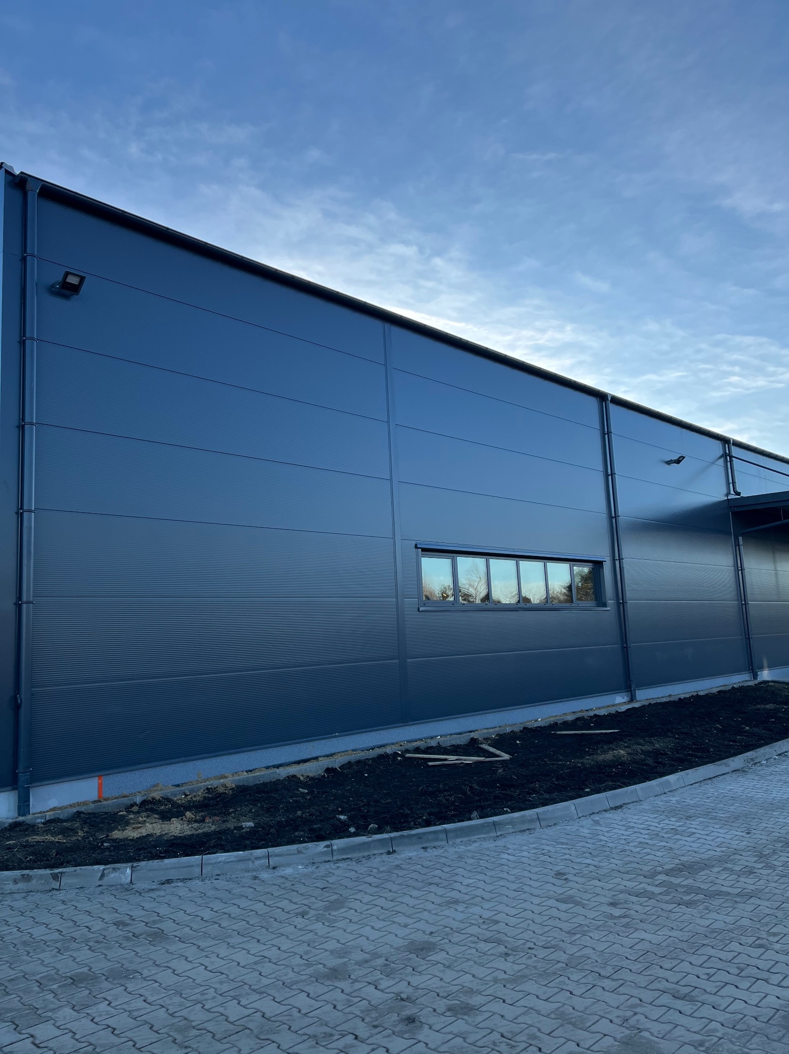 Elewacja hali przemysłowej z oknem i lampą zewnętrzną, widok z poziomu chodnika z kostki brukowej, błękitne niebo w tle. Minimalistyczny design.