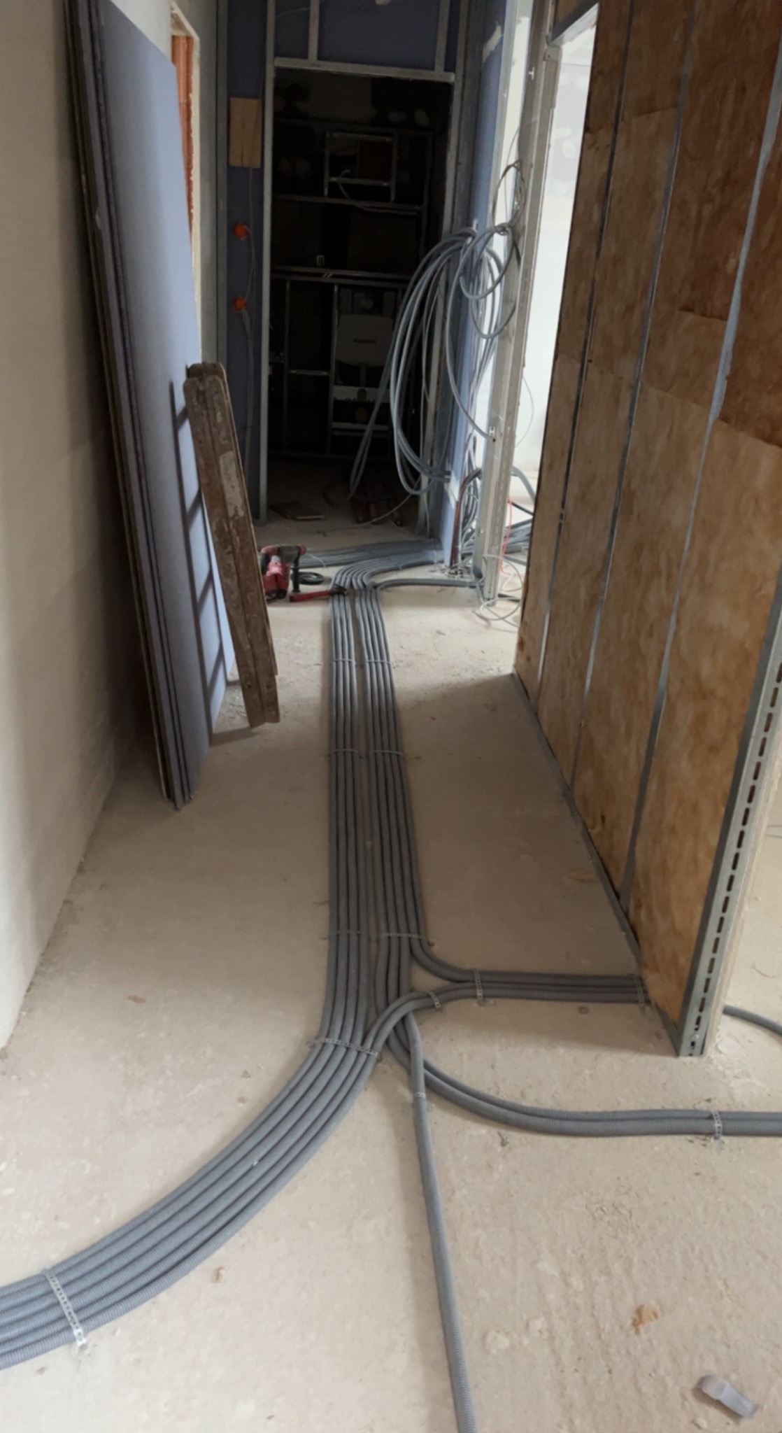 Szare rury instalacyjne ułożone równolegle na podłodze w surowym wnętrzu, widoczne narzędzia i elementy konstrukcyjne ścian. Etap prac instalacyjnych.