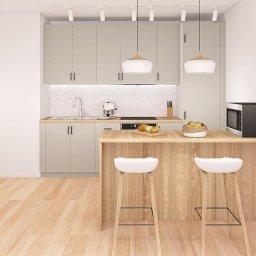 Wnętrze z sensem - Jasna kuchnia z drewnianym blatem i białymi hokerami. Nad blatem wiszą dwie lampy. Na blacie owoce i mikrofalówka. W tle białe płytki na ścianie.